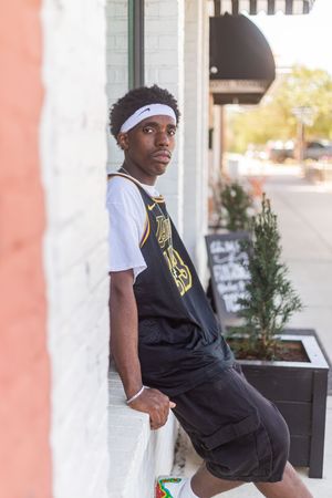 Young Black man in Nike headband and basketball jersey leans against white brick wall outside urban storefront.