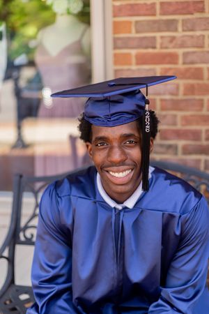 Smiling young man in blue graduation cap and gown seated outdoors near brick wall celebrating academic achievement.