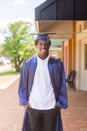 Young man smiling in blue graduation cap and gown standing outdoors on a brick walkway.