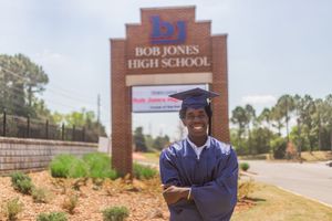 Graduate in cap and gown poses proudly in front of Bob Jones High School sign on a sunny day.