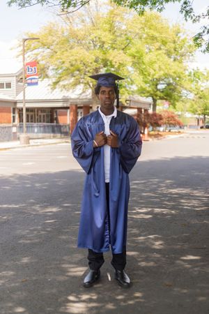 Young man in blue graduation cap and gown standing outdoors on a sunny day near trees and a brick building.