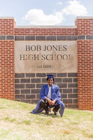 Graduate in blue cap and gown poses proudly in front of Bob Jones High School established 1974 sign.