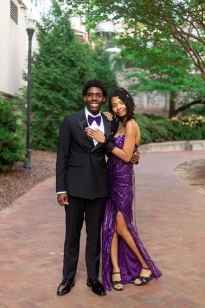 A couple dressed in prom attire, man in black tuxedo and woman in purple sequin gown, posing outdoors.