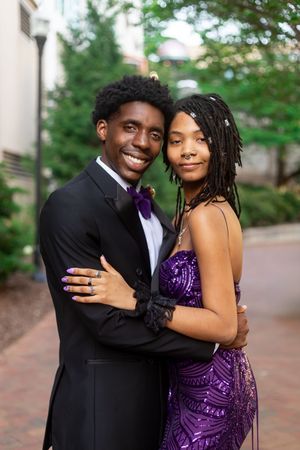 A smiling couple dressed in prom attire, man in black tuxedo and woman in purple sequin gown, posing outdoors.