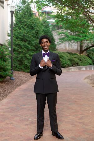Young man in black tuxedo with purple bow tie smiling outdoors on brick path surrounded by lush green hedges.