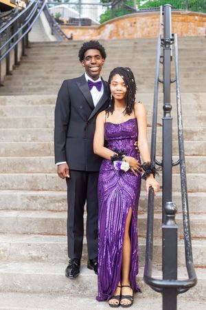 A young couple dressed in prom attire, posing on stone steps, with the man in a tuxedo and woman in a purple sequin gown.