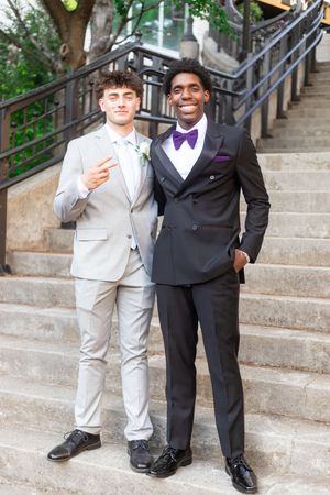 Two young men in formal suits pose on outdoor steps, one in gray with boutonniere, one in black tuxedo with purple bow tie.