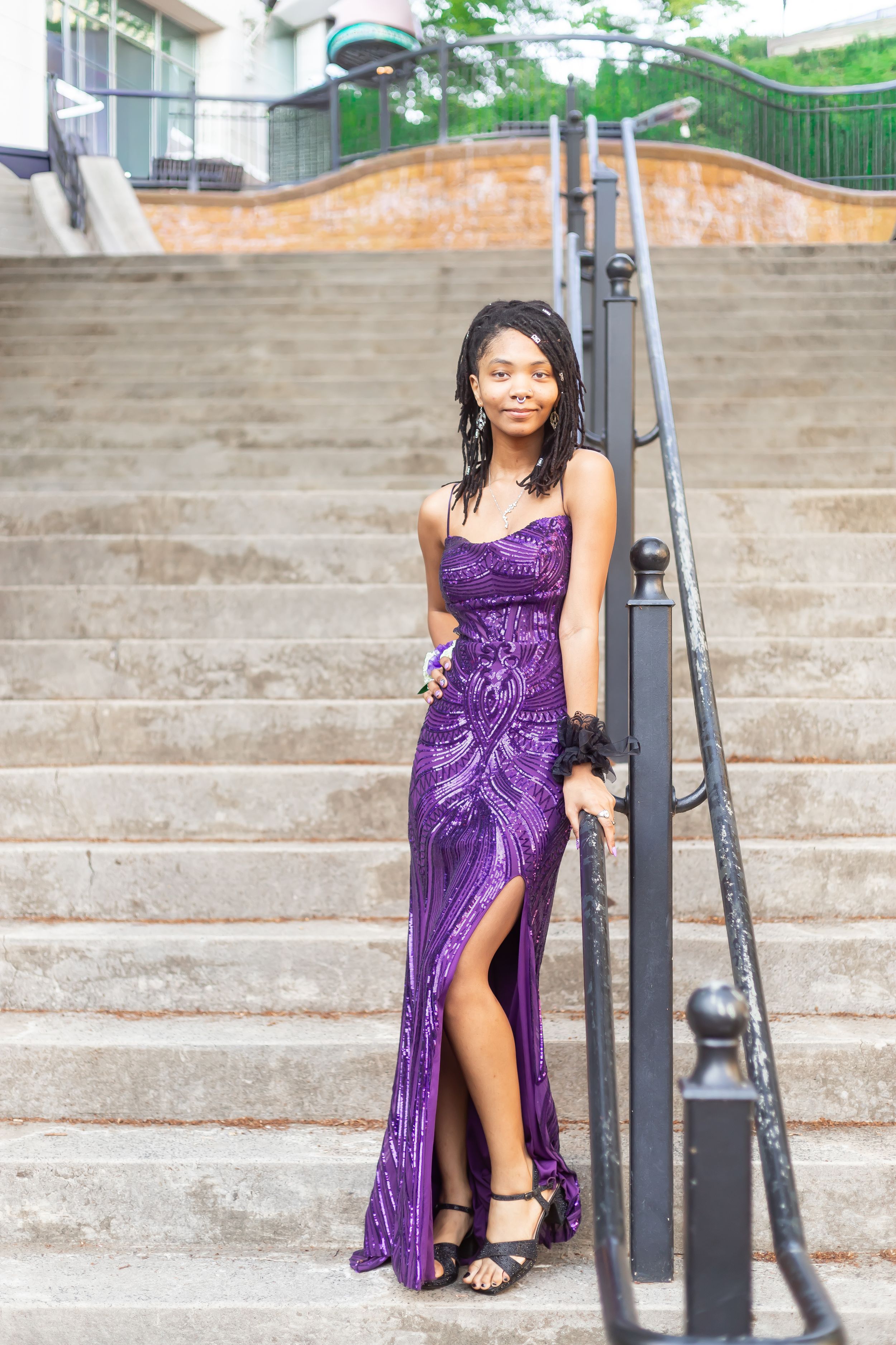 Young woman in a sparkling purple sequin gown with slit poses on stone steps, wearing black heels and wrist corsage.