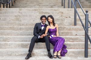 A young couple in formal prom attire, man in black tuxedo and woman in purple gown, posing on stone steps.