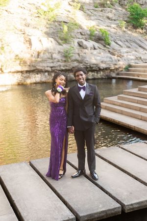 A couple dressed in prom attire poses on a wooden bridge, she in a purple sequin gown, he in a black tuxedo.