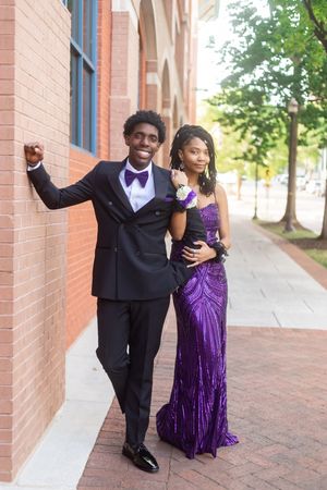 A young couple dressed in prom attire poses outdoors, he in a black tuxedo and she in a purple sequin gown.