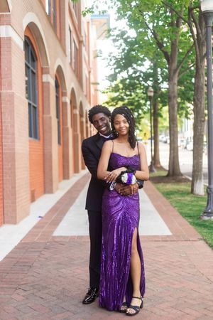 A young couple dressed in prom attire, with the woman in a purple sequin gown and man in a black tuxedo, posing outdoors.