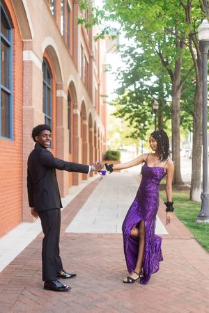 A young couple in prom wear hold hands outdoors, she in a purple sequin dress, he in a black suit.