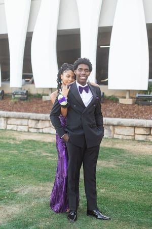 A young couple dressed in prom attire, man in black tuxedo and woman in purple gown, posing outdoors.