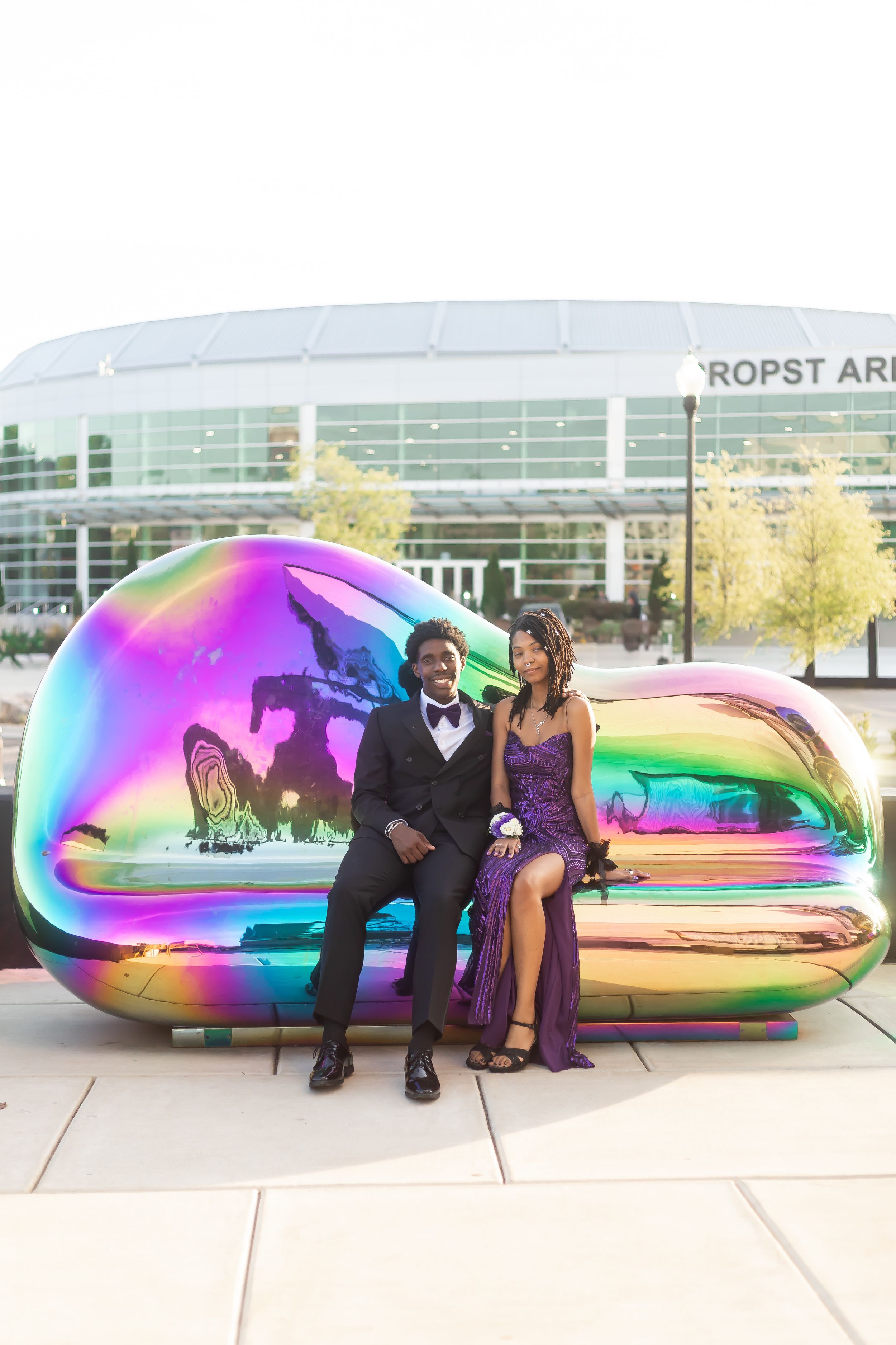 A prom couple in formal wear sits on a colorful iridescent metallic sculpture outside Propst Arena.