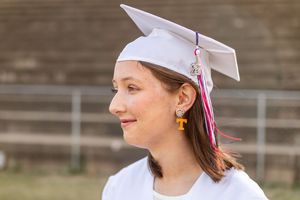 Graduate in white cap and gown smiles during outdoor graduation ceremony celebration.