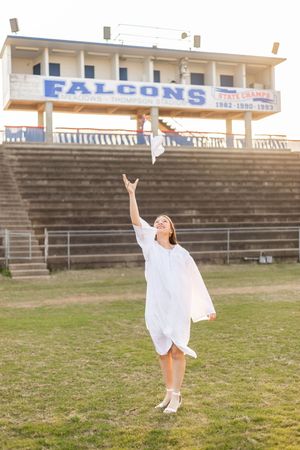 A person in a flowing white dress twirls joyfully in front of stadium bleachers on a grassy field.