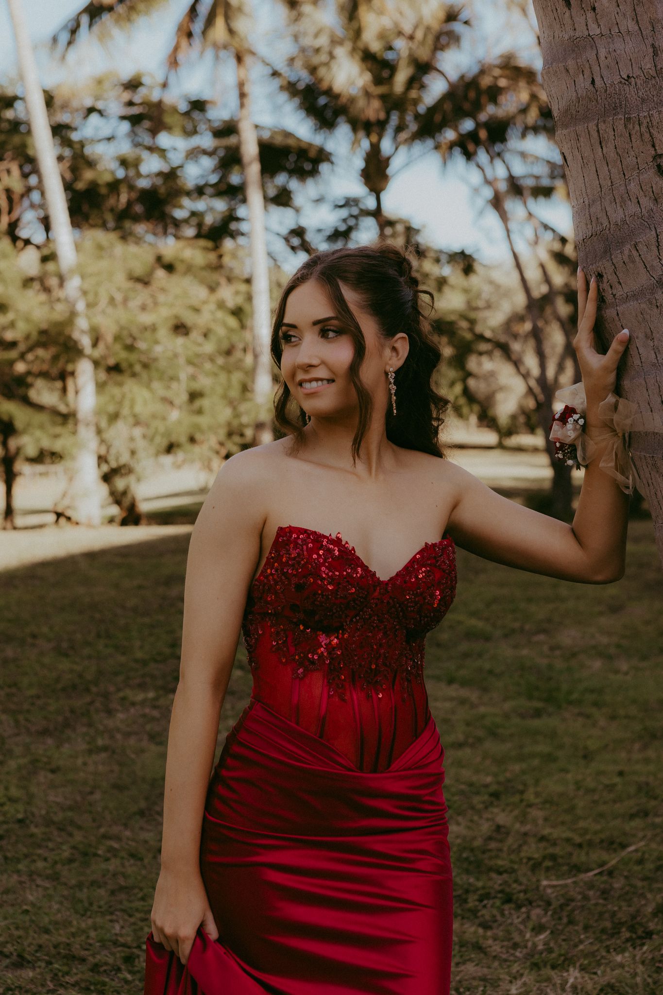 Young woman in red sequin strapless gown posing by palm tree at prom.