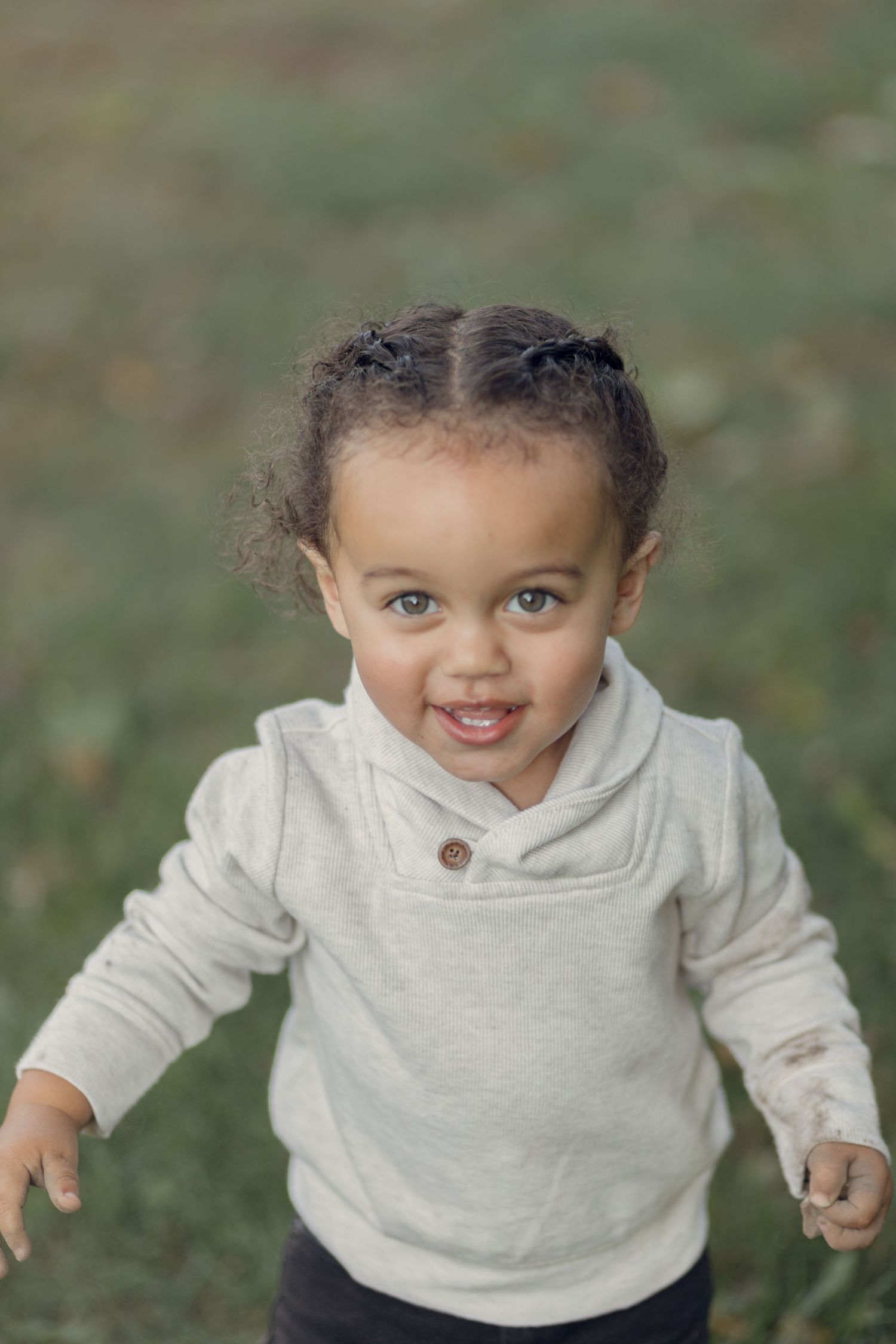 Child in beige sweater smiling brightly outdoors.