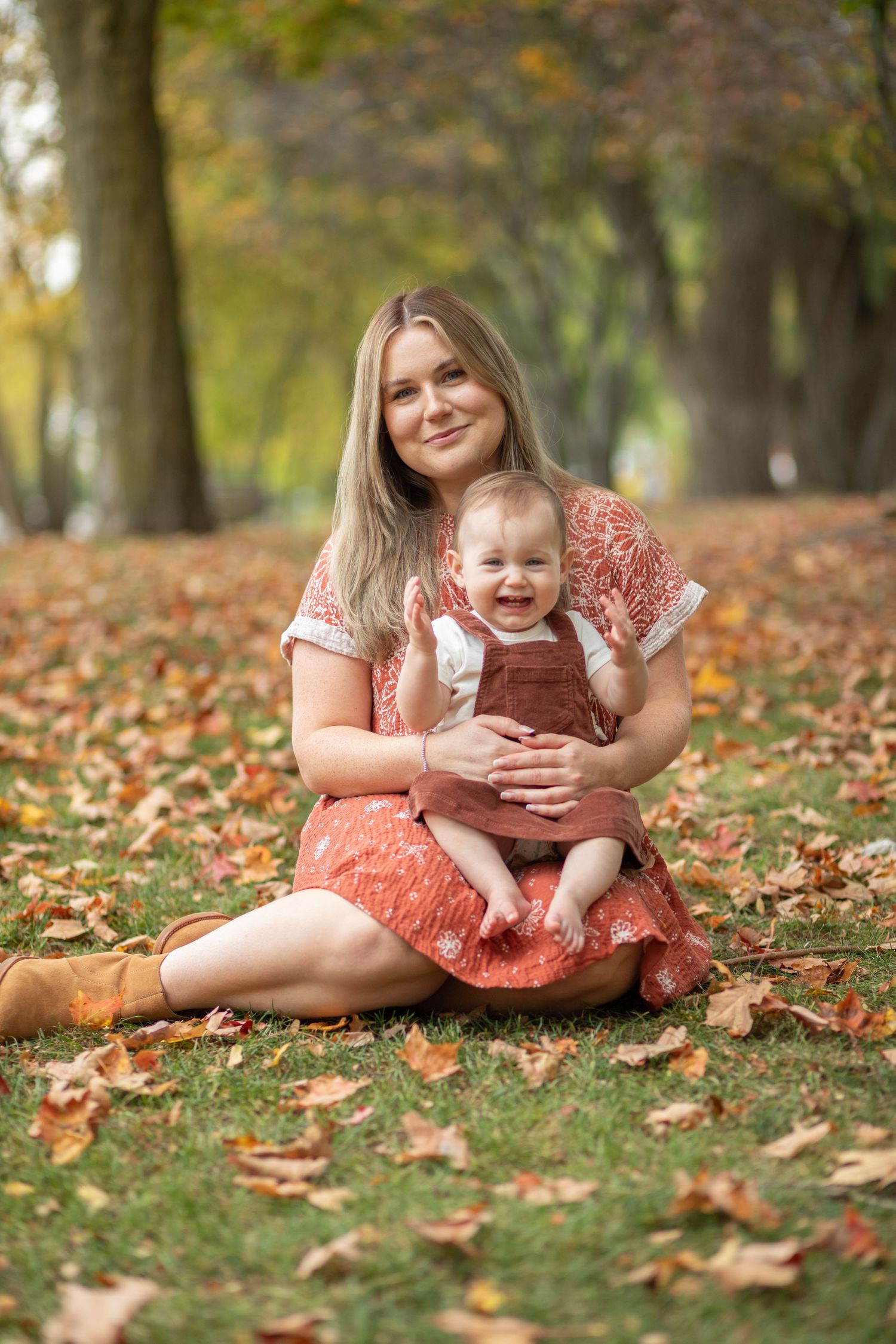 Mother and baby sitting on grass surrounded by fallen autumn leaves.