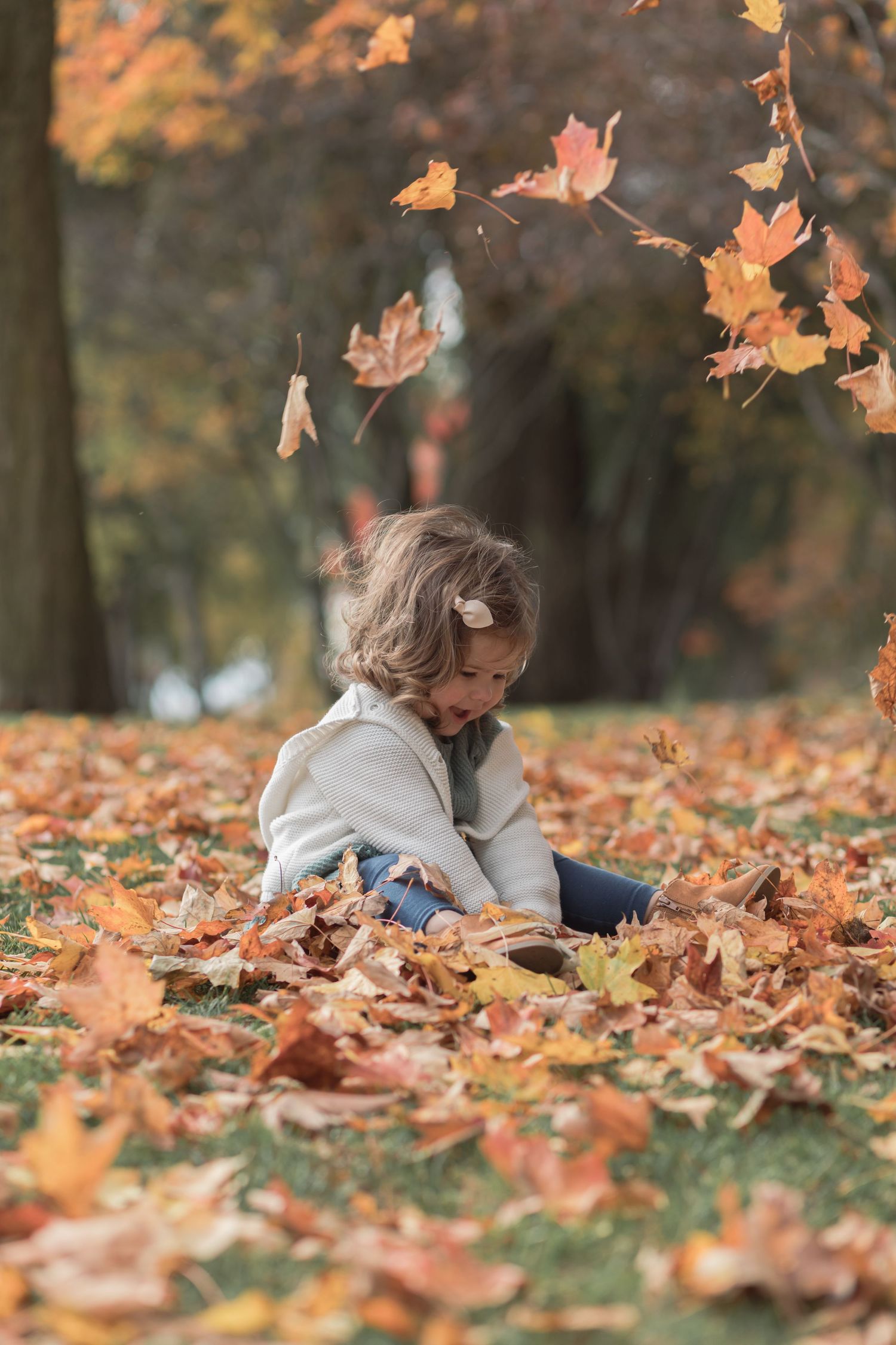 Child sitting in fallen autumn leaves as maple leaves float through the air.