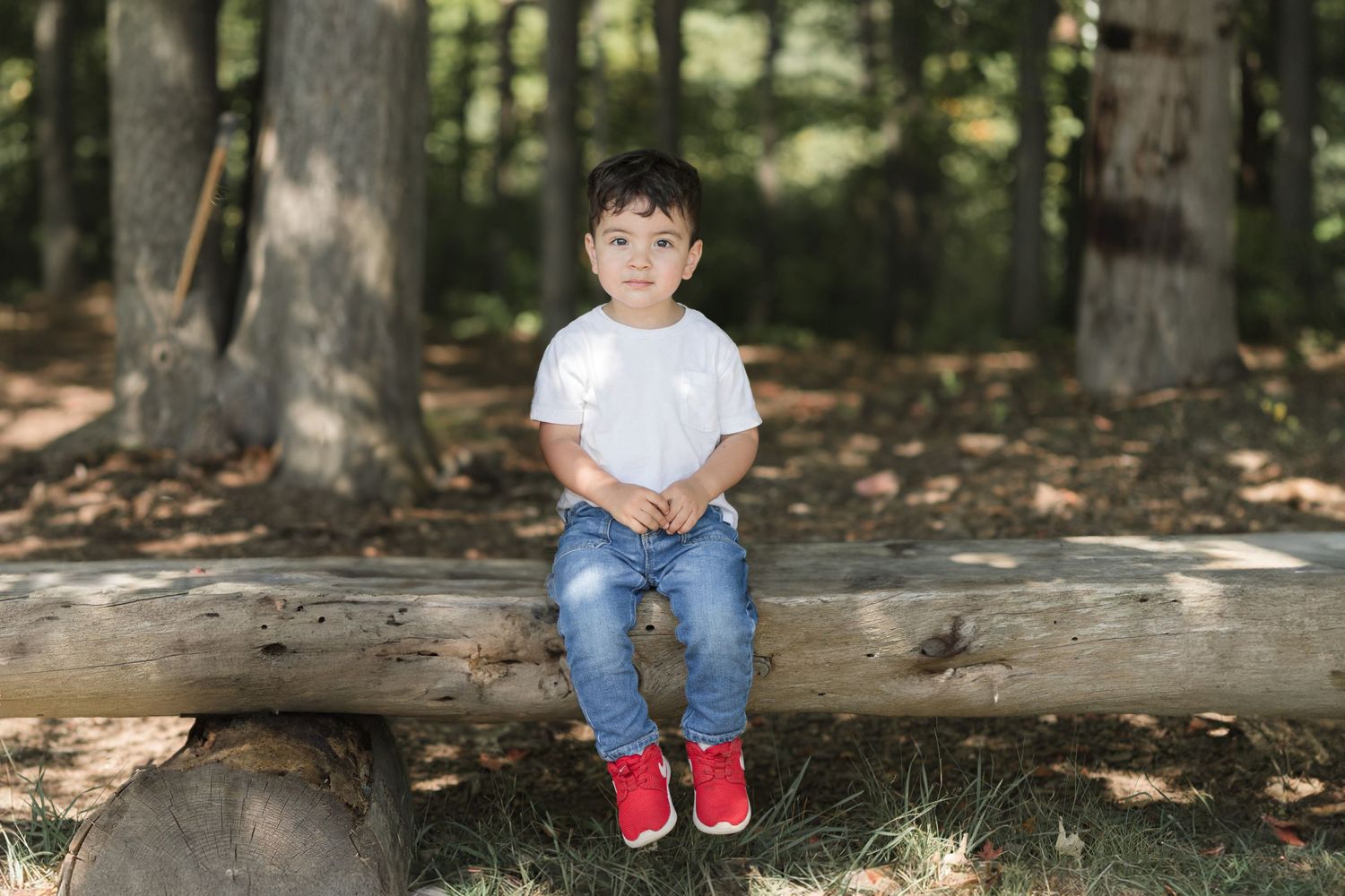 A child in a white t-shirt and jeans sits on a fallen log in a forest setting wearing bright red shoes.