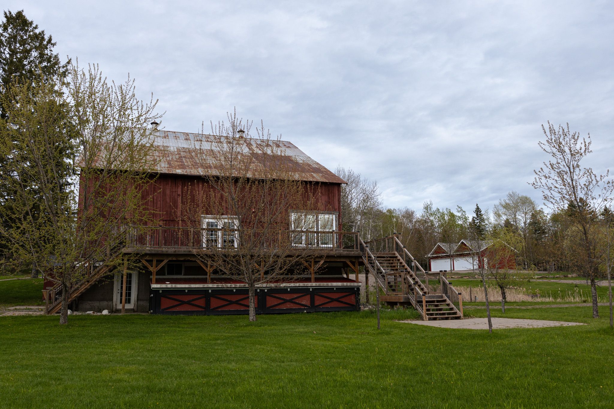 Rustic red barn with wooden stairs and deck set on green lawn under cloudy sky.