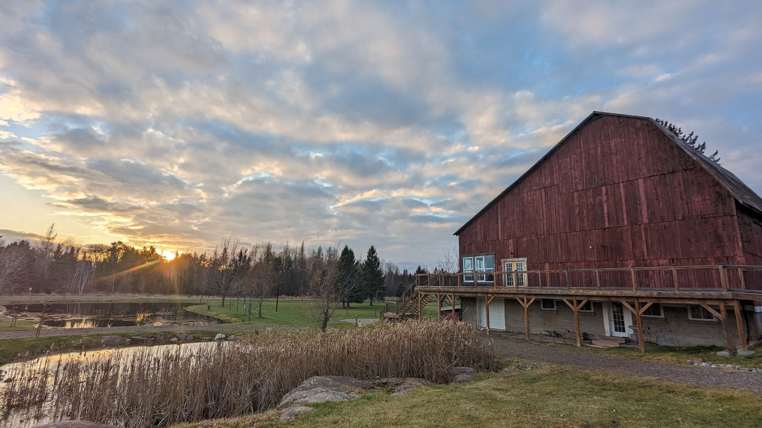 A weathered red barn sits beside a pond at sunset with dramatic clouds and evergreen trees in the background.