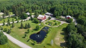 Aerial view of a rural property with red barn, manicured lawn, decorative pond and surrounding forest landscape.