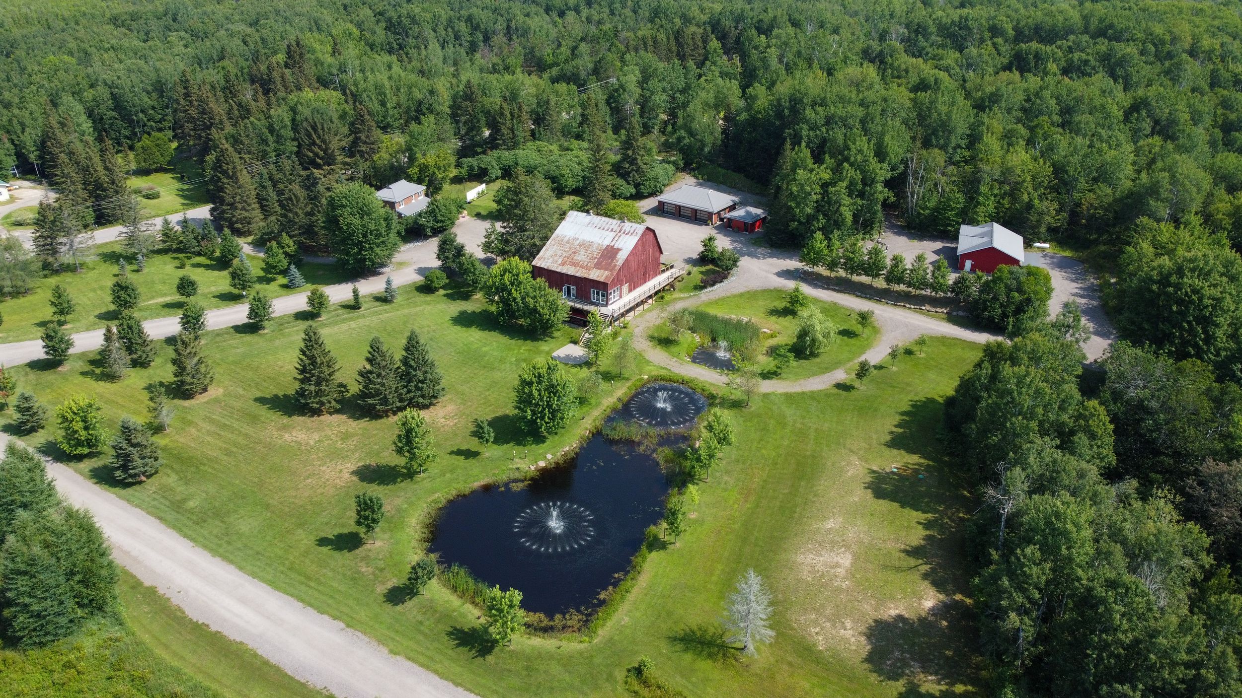 Aerial view of a rural property with red barn, manicured lawn, decorative pond and surrounding forest landscape.
