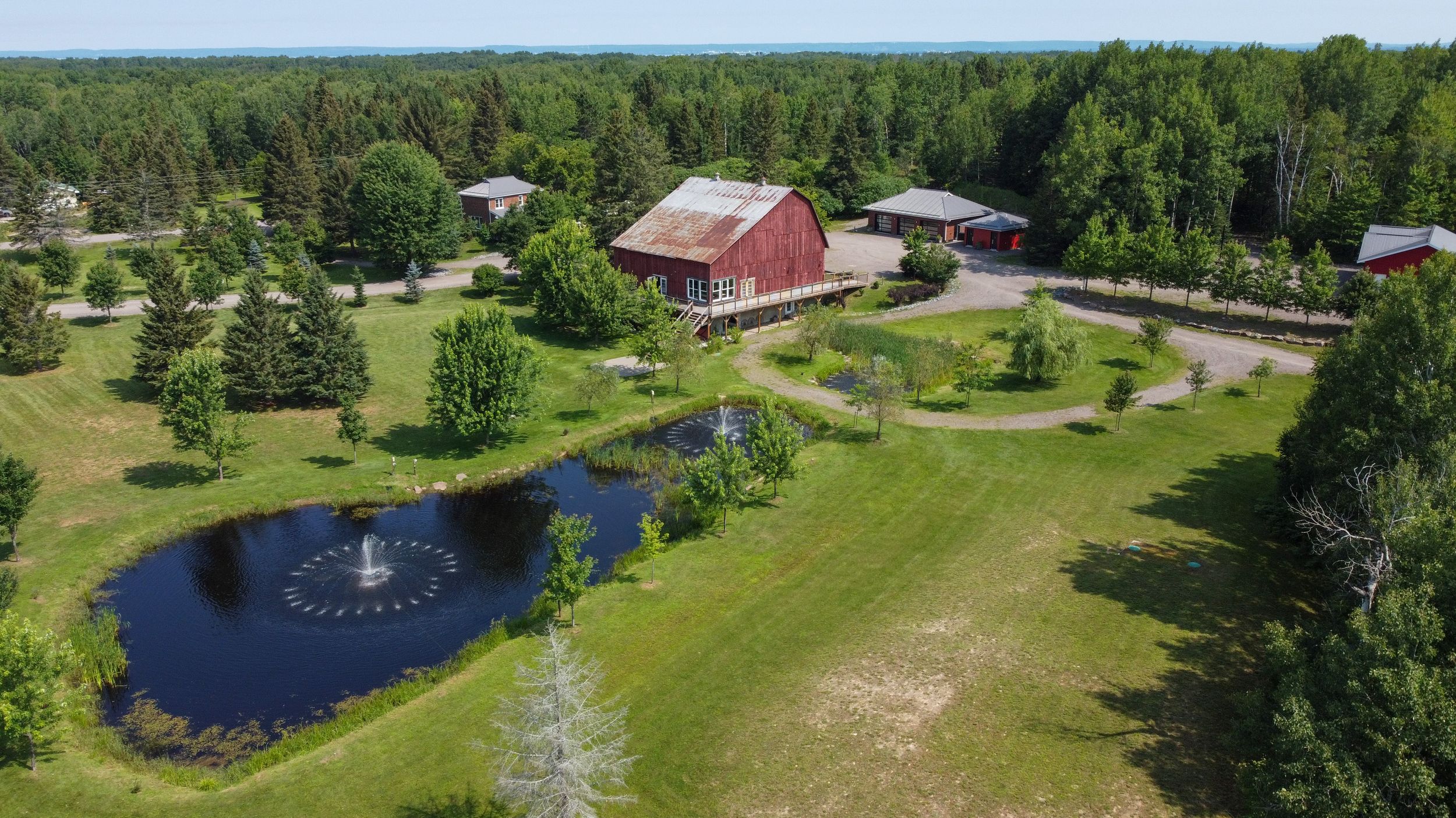 Aerial view of a rustic red barn with a scenic pond featuring a fountain surrounded by manicured lawns and trees.