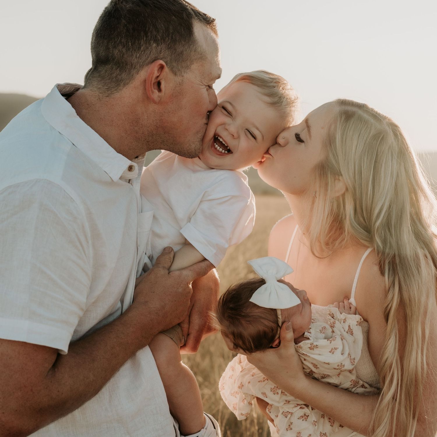 Happy family with parents kissing laughing toddler while mother holds newborn baby in golden sunlight.