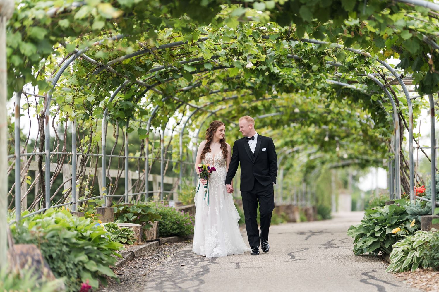 Wedding couple strolling through garden path surrounded by green foliage and natural light.