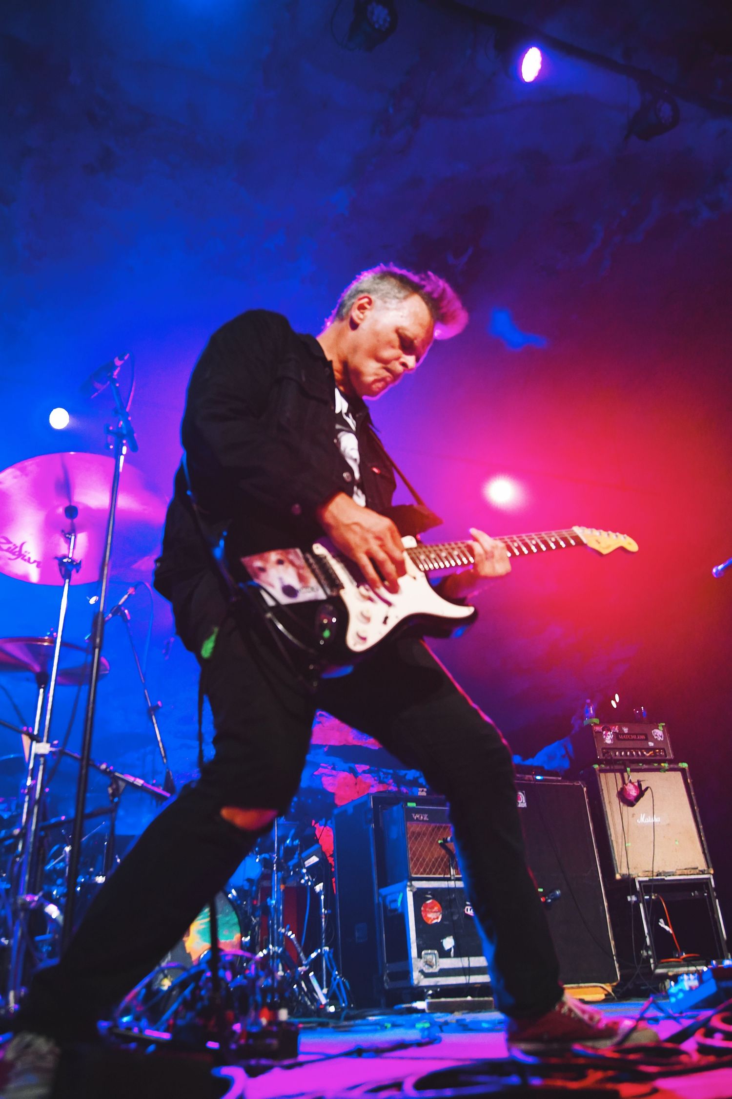 Guitarist performs energetically on stage with vibrant blue and pink lighting creating a dynamic concert atmosphere.