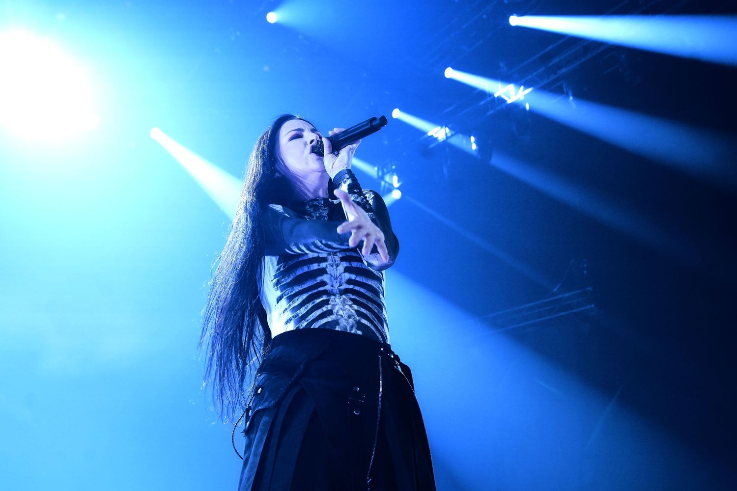Female singer in skeleton corset performs on blue-lit stage, holding microphone with dramatic stage lighting behind her.