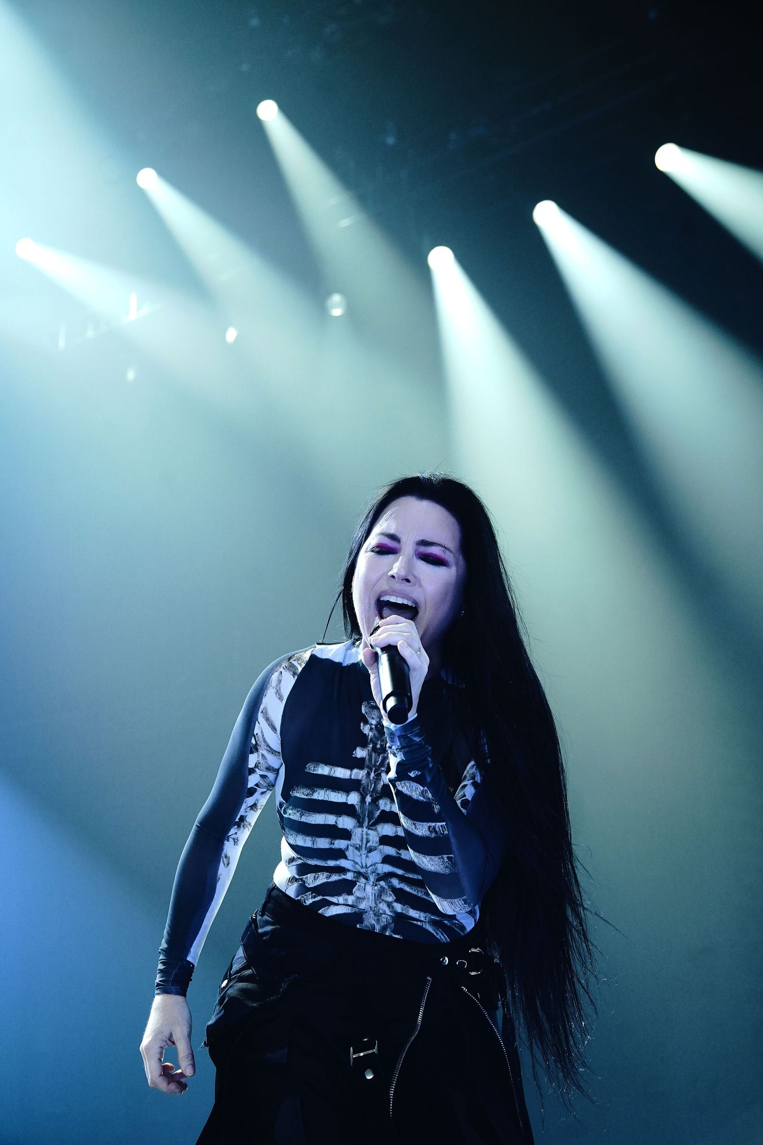Female rock singer performing on stage in skeleton top, long black hair, under dramatic blue spotlights.