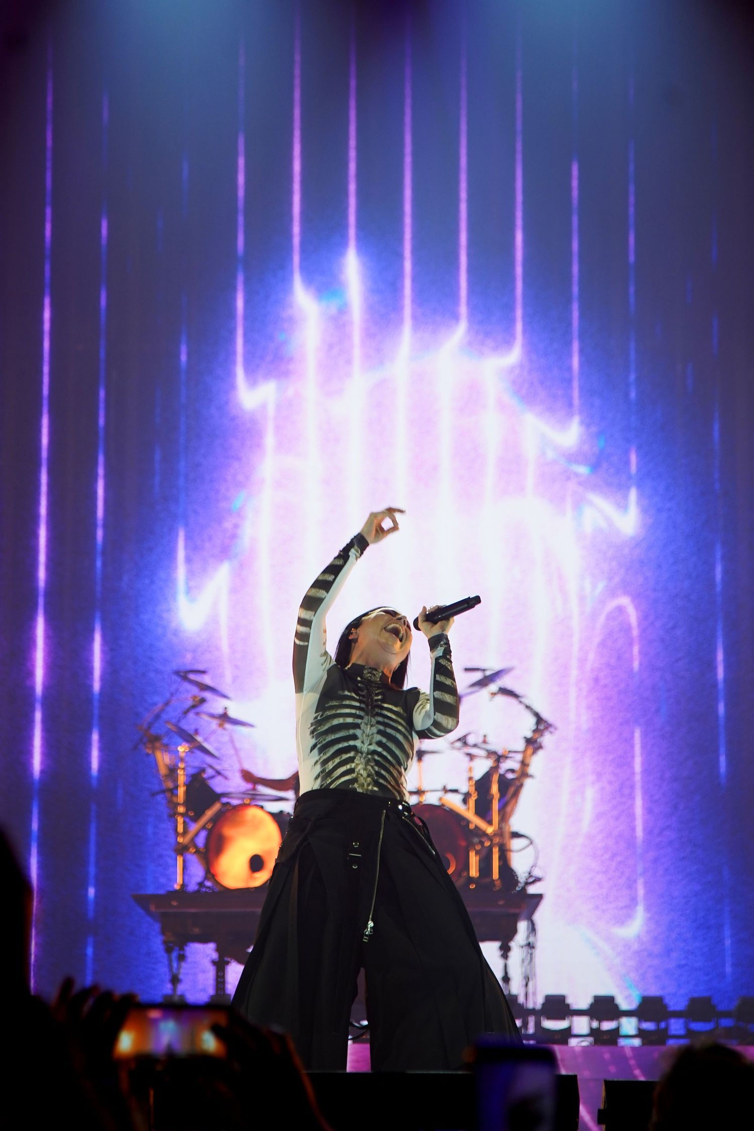 Female singer performing on stage with dramatic purple lighting, wearing striped bodysuit and wide-leg pants, holding microphone up.