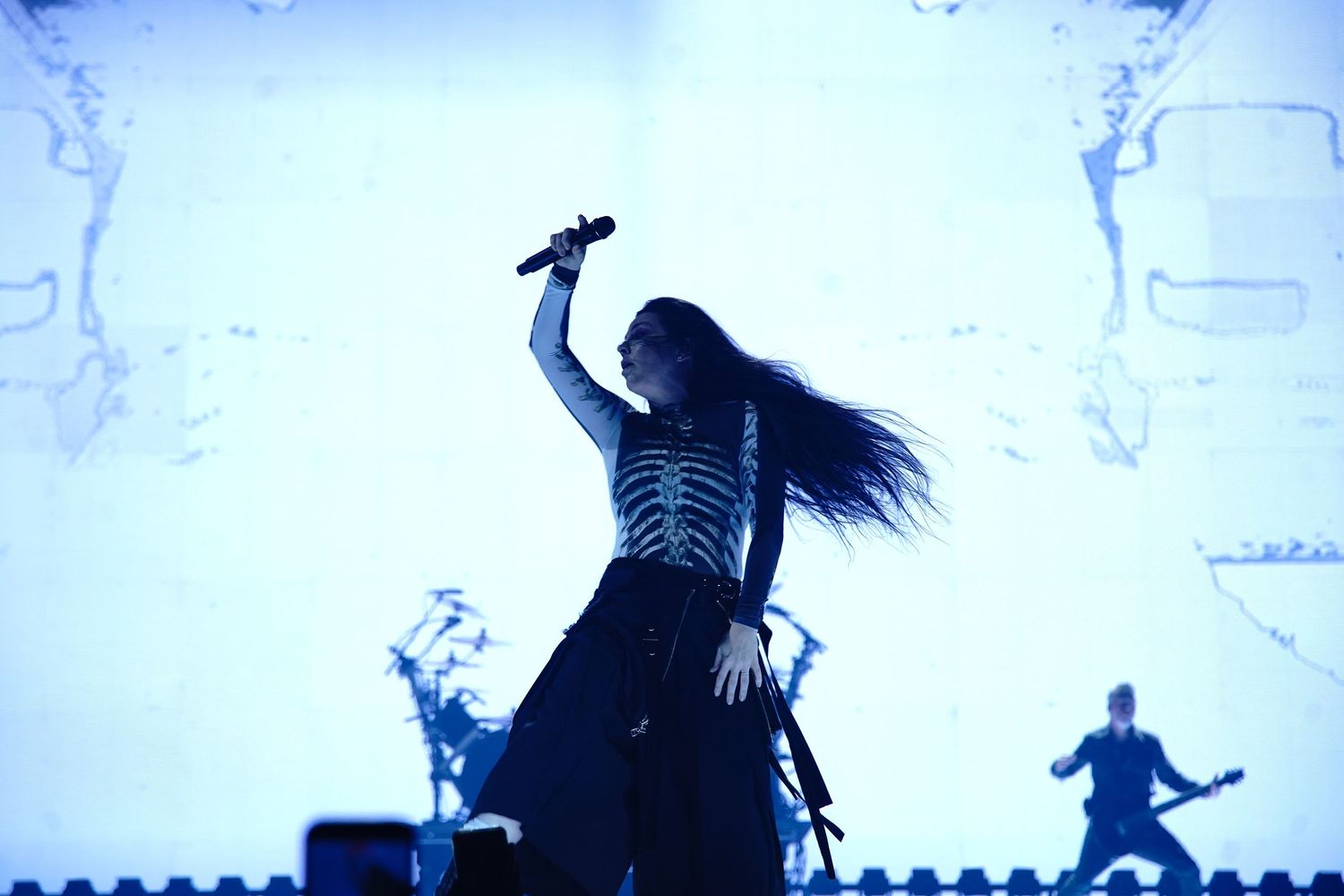 Female rock singer performing on stage with blue lighting, raising microphone dramatically with long flowing hair.