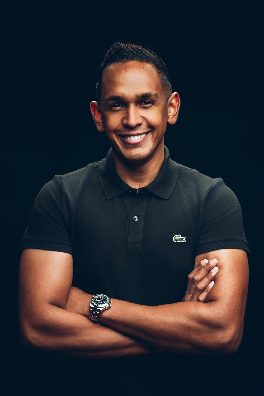 Professional portrait series against dark background showing confident pose in Lacoste polo shirt with crossed arms.