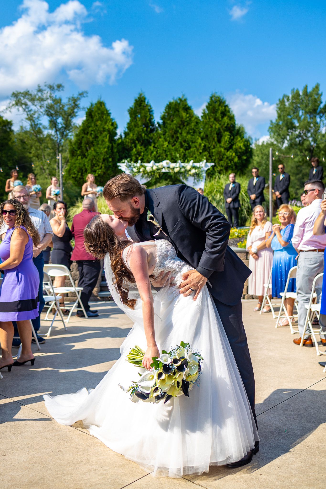 Wedding couple performs romantic dip kiss on outdoor patio during sunny summer ceremony.