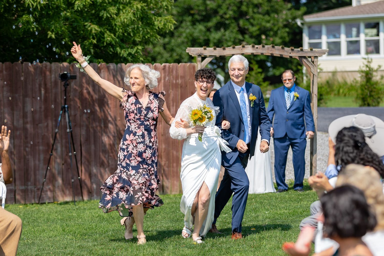 Wedding guests celebrate joyfully while running across a grassy lawn.
