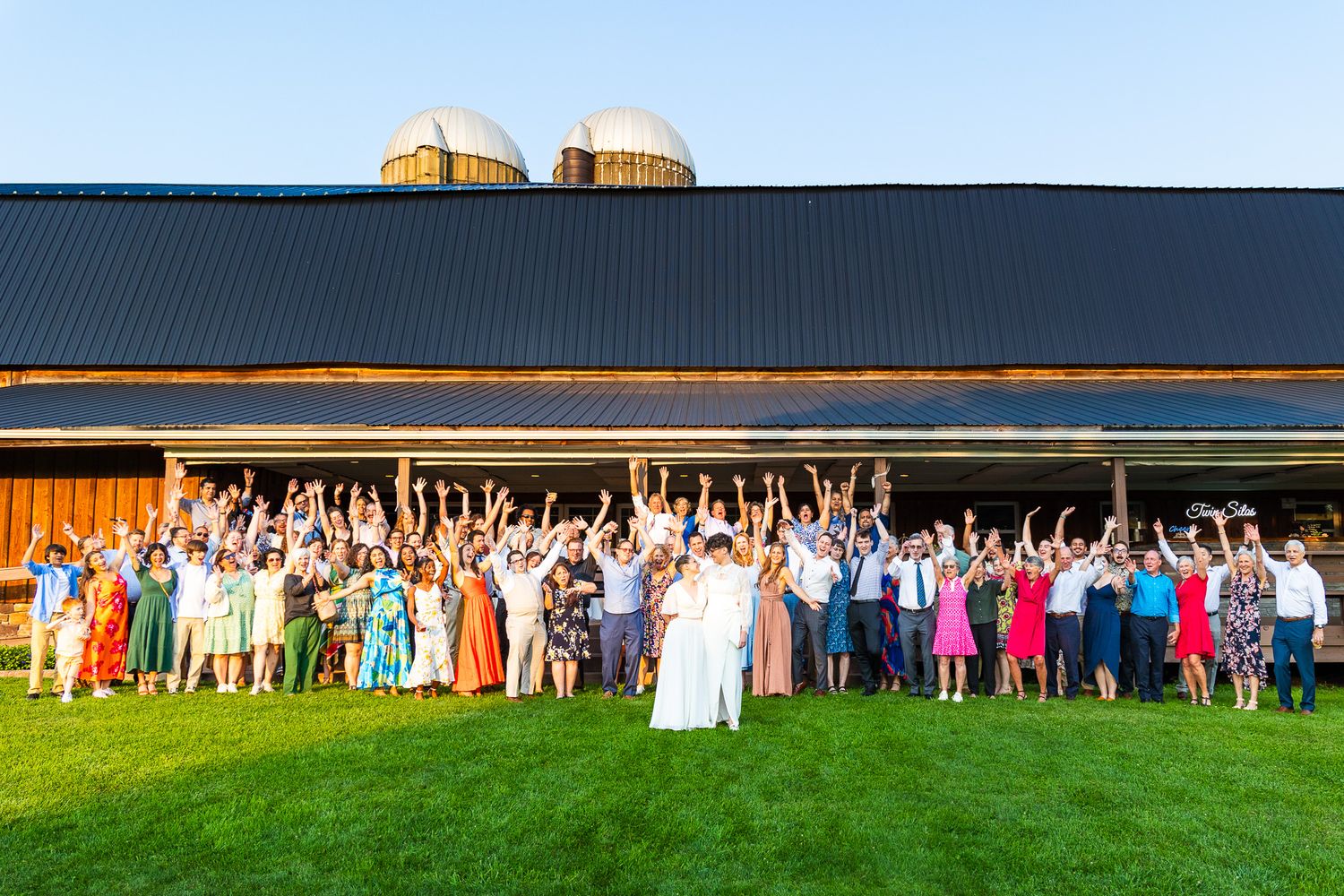 Large group wedding photo with guests lined up in front of modern barn building.