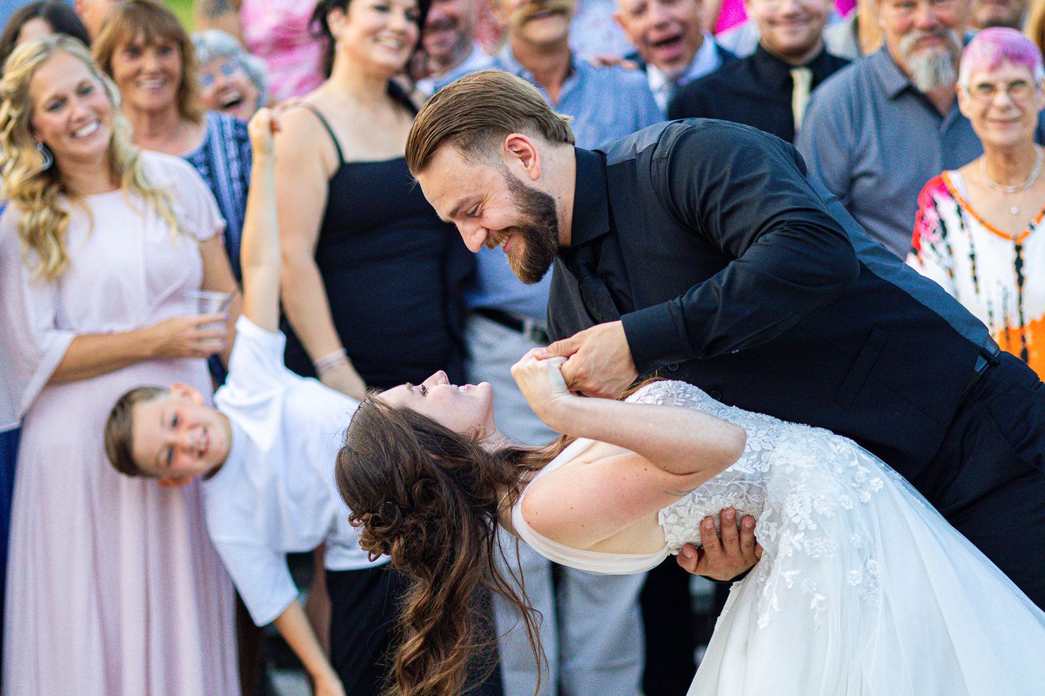 Dramatic dip kiss at outdoor wedding with guests cheering in background.