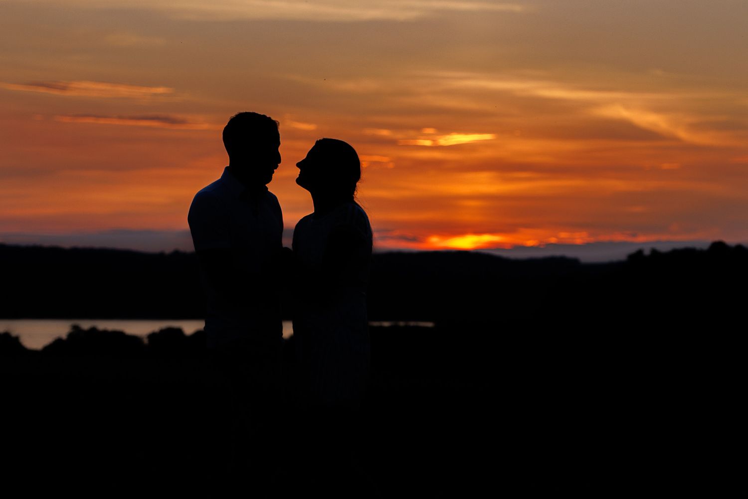 Silhouetted figures stand against a vibrant orange sunset sky with dark landscape in foreground.