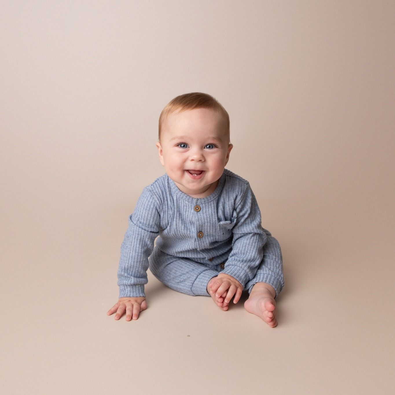 Happy baby boy in blue striped romper sitting and smiling on a beige studio background.