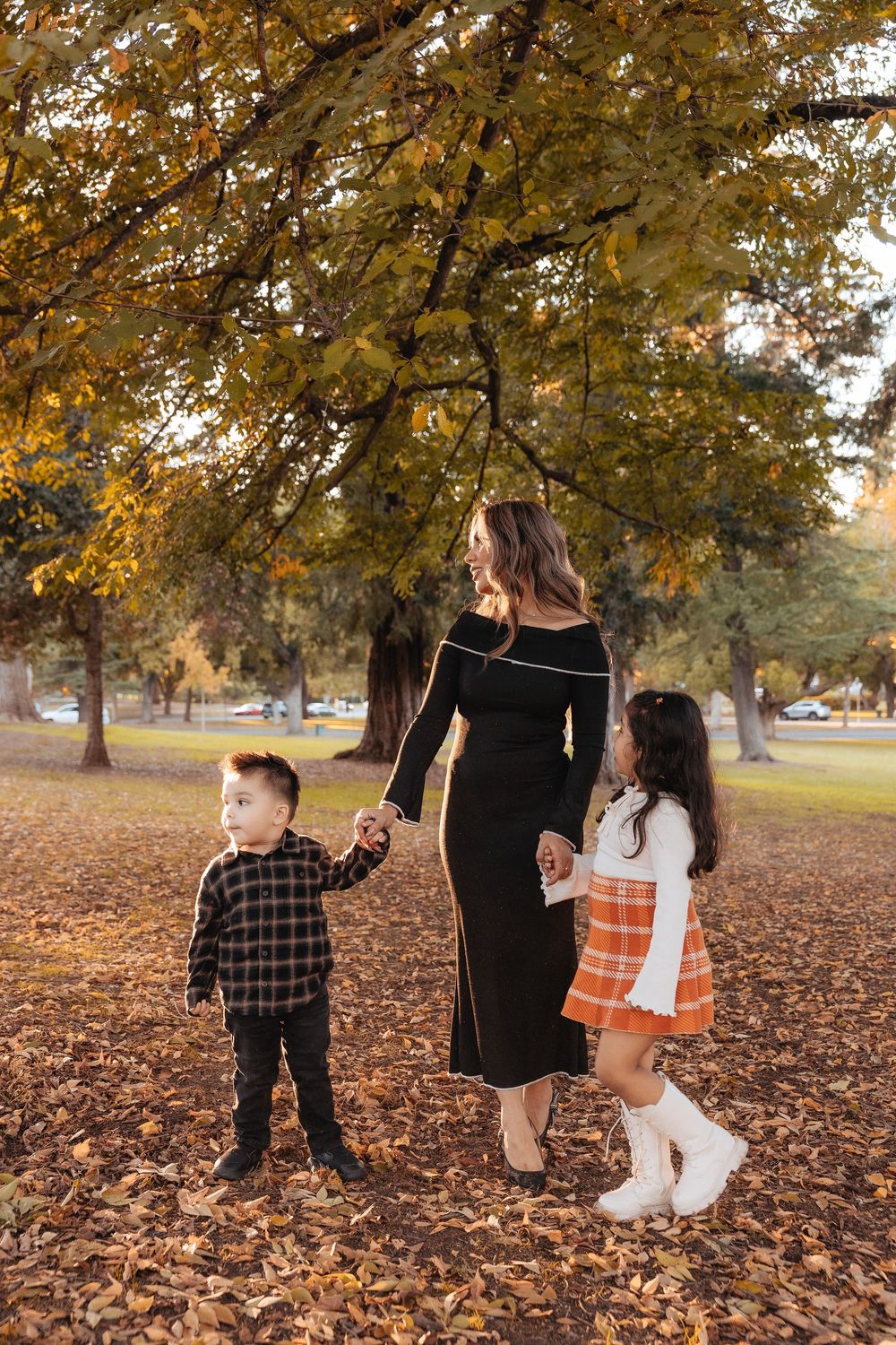 A family walks through fallen autumn leaves in a park during golden hour as trees display warm fall colors.
