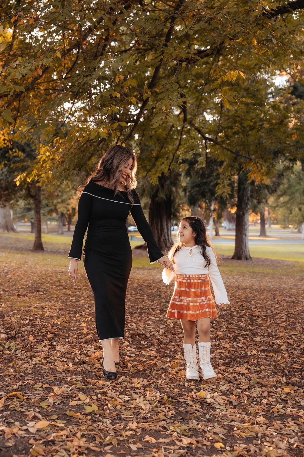 Two people walk hand in hand through a park covered in fallen autumn leaves on a crisp fall day.