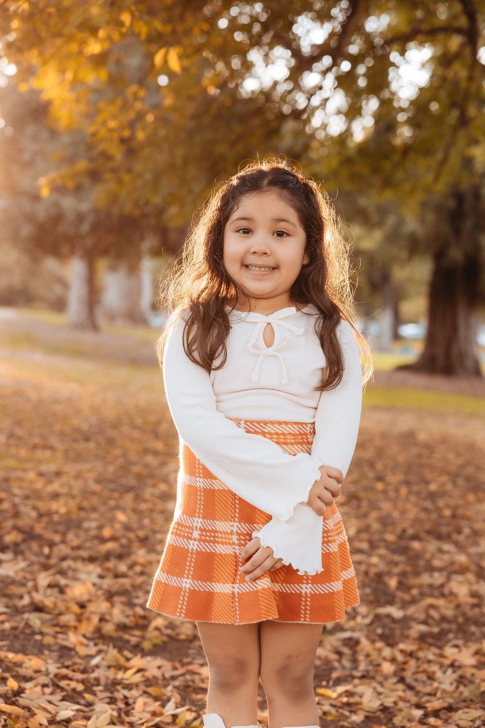 Child wearing white and orange dress poses in autumn leaves during golden hour sunset.