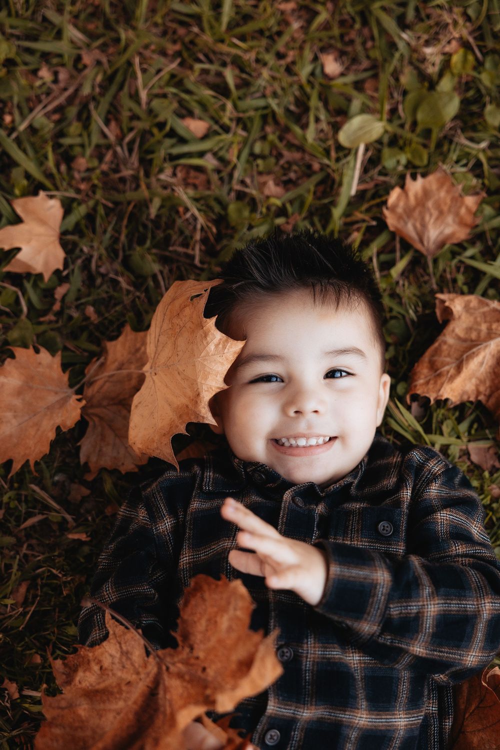 A child lies on autumn leaves wearing a plaid flannel shirt while smiling at the camera.