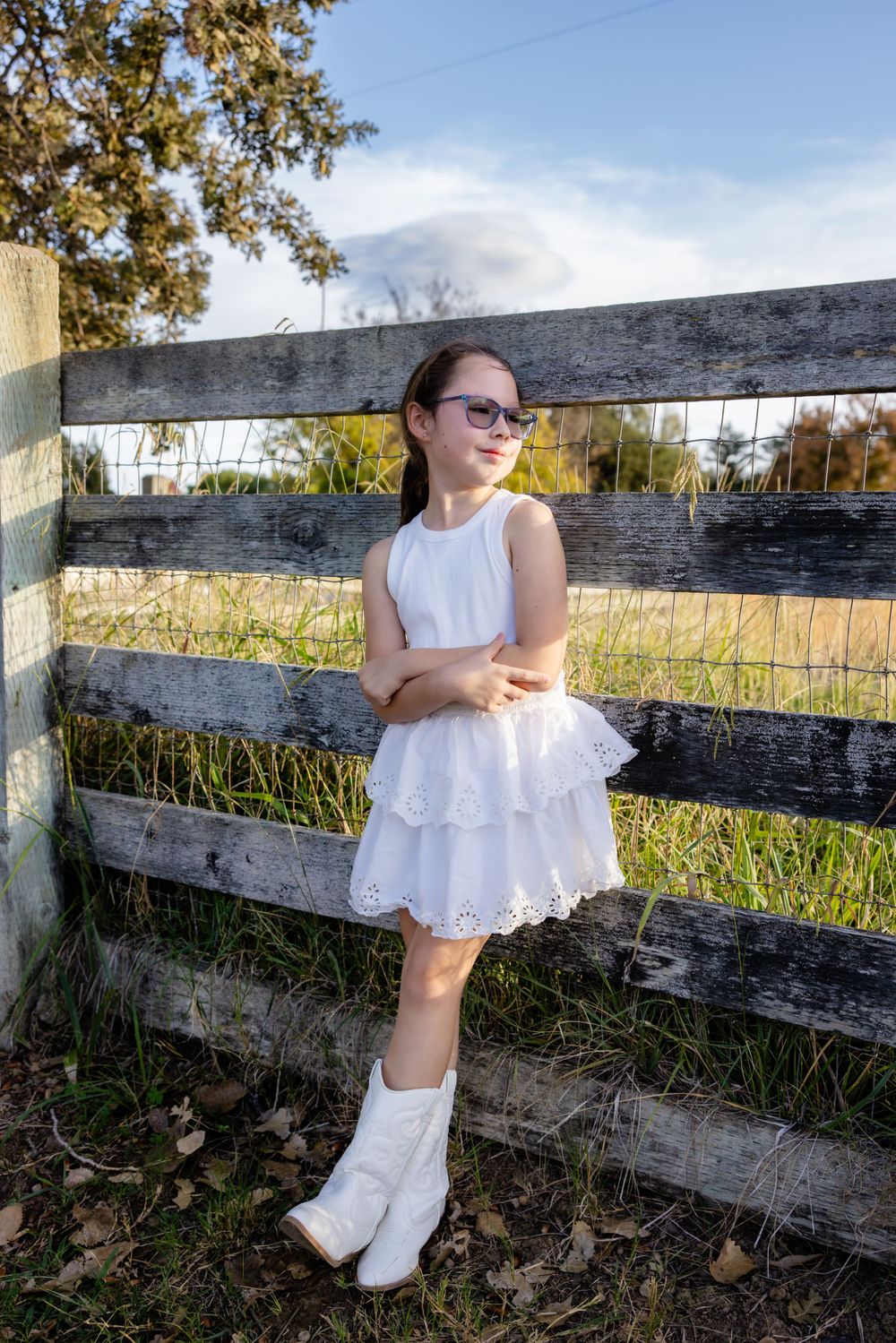 Young person in white dress poses by rustic wooden fence in countryside setting.